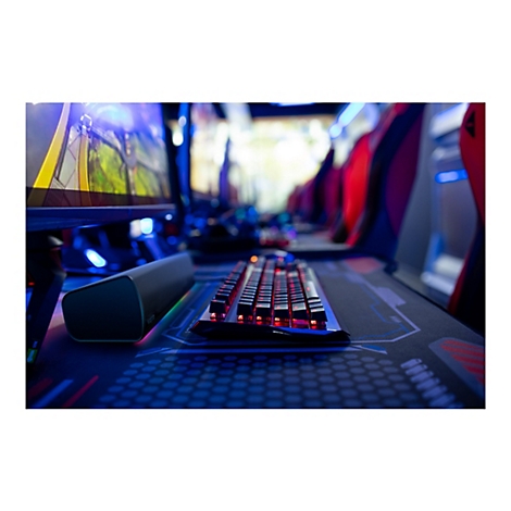 Eine Nahaufnahme einer beleuchteten Gaming-Tastatur, die sich auf einem Gaming-Schreibtisch befindet. Im Hintergrund sind Monitore und eine Gaming-Umgebung zu sehen.