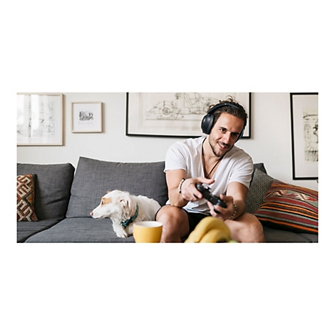 Ein Mann spielt mit KopfhÃ¶rern und Controller auf einer Couch. Ein Hund und eine Tasse sind auch zu sehen.