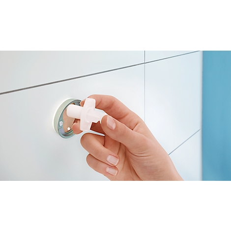 Une main tient un bouchon blanc en forme de croix. En arrière-plan, un mur blanc avec un support rond.