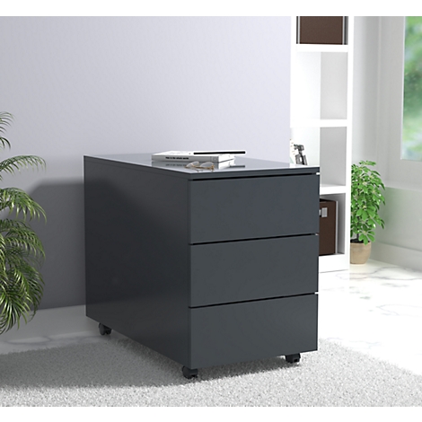 Dark gray mobile pedestal with three drawers. A book and glasses lie on top. In the background: shelf, plant.