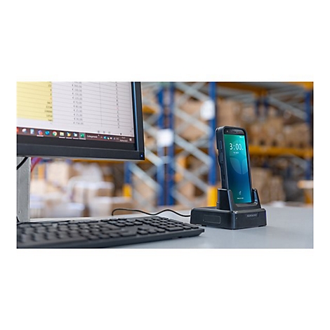 Ein Smartphone in einer Ladestation mit Uhrzeit 3:00, am Schreibtisch neben Monitor und Tastatur. Im Hintergrund ist ein Lager erkennbar.