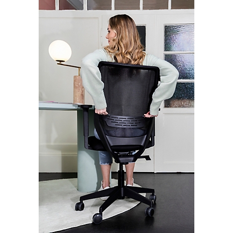 Eine Frau sitzt auf einem schwarzen Bürostuhl, ihre Hände am Stuhl. Der Hintergrund ist unscharf, mit einem Schreibtisch und einer Lampe.