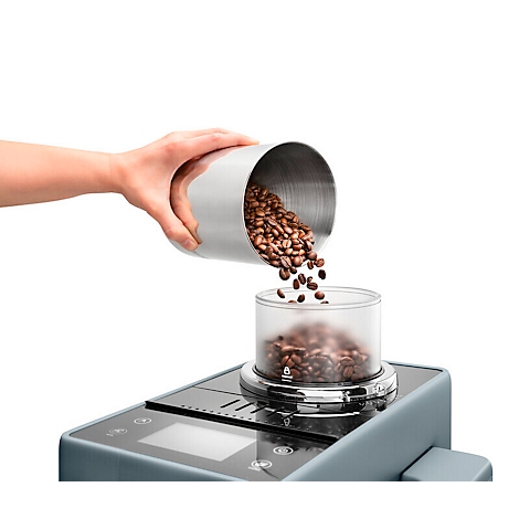 Hand schüttet Kaffeebohnen aus einem silbernen Behälter in eine transparente Kammer.