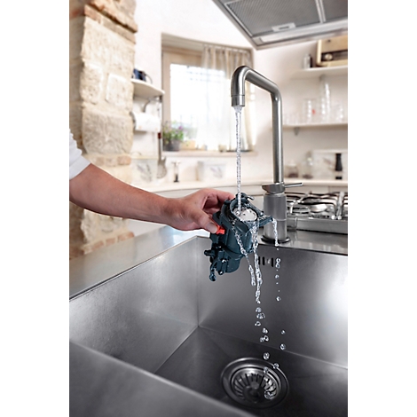 Een persoon spoelt een donker apparaat af onder stromend water in een gootsteen. Op de achtergrond is een keuken te zien.