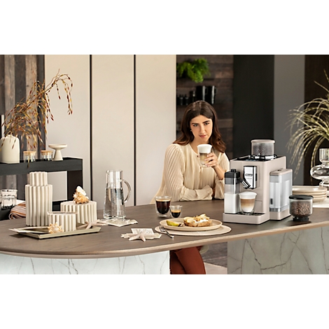 Een vrouw met een cappuccino zit aan de bar naast een koffiemachine. Koffie, gebak en decoraties staan ook op tafel.