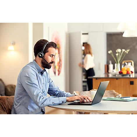A man with a headset types on a laptop at a table. A woman is in the background in the kitchen.