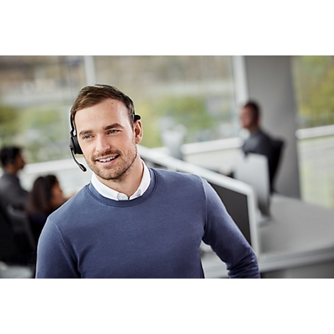 A smiling man wearing a headset in an office. Other people and screens are visible in the background.