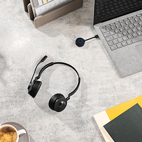 Black headset lies next to laptop, coffee cup and notebooks on a light desk.