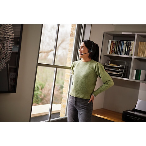 Woman with headset looks out of a window in an office. She smiles relaxed, arm on hip.