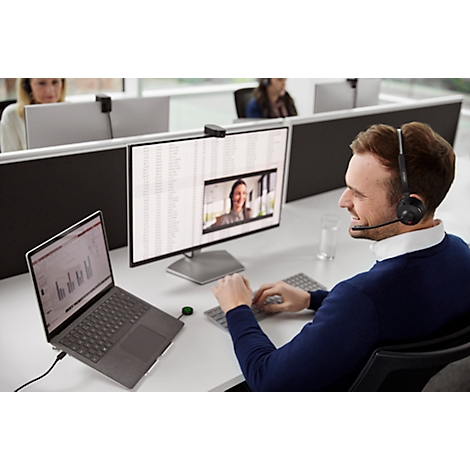 A man wearing a headset at a desk, in a video call, a woman can be seen on the monitor, other people in the background.