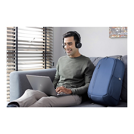 Ein Mann mit Headset sitzt auf einem Sofa und arbeitet an einem Laptop. Neben ihm steht ein blauer Rucksack.
