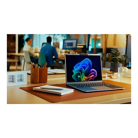 Ein Laptop mit Windows-Hintergrund steht auf einem Schreibtisch, im Hintergrund arbeiten zwei Personen.