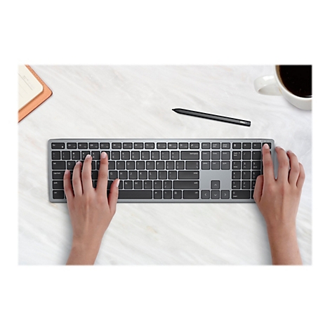 Gros plan d'une personne plaçant ses mains sur un clavier d'ordinateur argenté, avec un stylet noir et une tasse de café en arrière-plan.