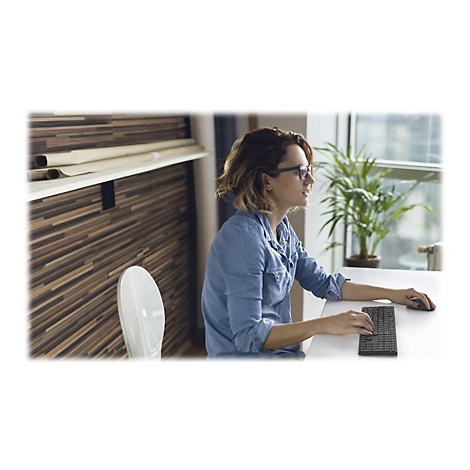 Eine Frau sitzt an einem Tisch und arbeitet am Computer. Sie trägt eine Jeansjacke und Brille.