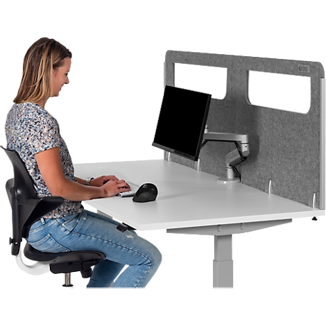 Woman sitting at a height-adjustable desk with a monitor, typing on a keyboard. Gray wall, black office chair, light wood.