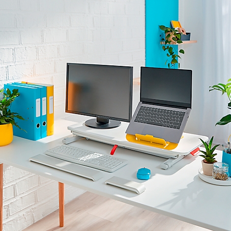A desk with a laptop, monitor, keyboard, and office supplies. Yellow laptop stand, plants, and yellow-blue folders.