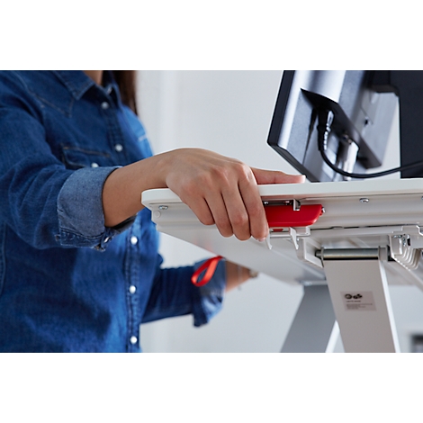 Person operating a height-adjustable desk; red lever, denim shirt, white tabletop, monitor in the background.