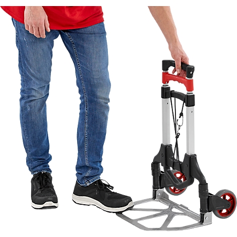 Personne avec un t-shirt rouge et un jean tenant un diable argenté avec des poignées et des roues rouges.