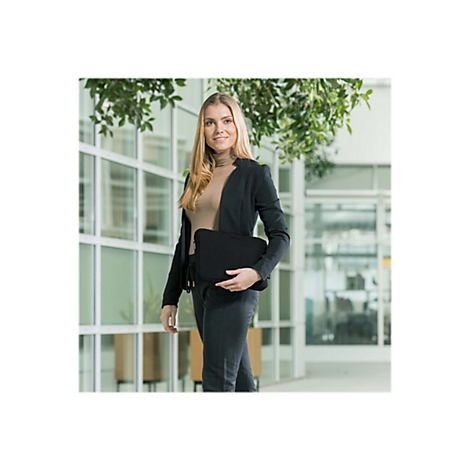 Eine Frau in schwarzem Blazer und Hose hält eine schwarze Laptoptasche. Sie steht in einem hellen Büro vor Fensterfront.