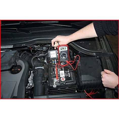Ein rotes Multimeter misst die Spannung an einer Autobatterie. Das Display zeigt 12,41 V an. Das Bild zeigt den Motorraum eines Autos.