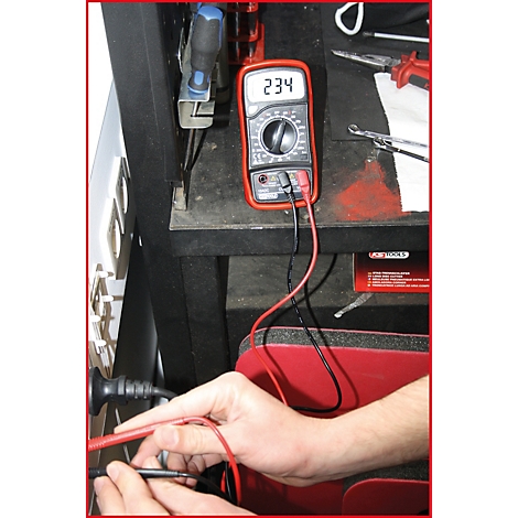 Ein Multimeter zeigt 234 an. Rote und schwarze Messleitungen sind angeschlossen, Hände halten weitere Leitungen. Werkbank.
