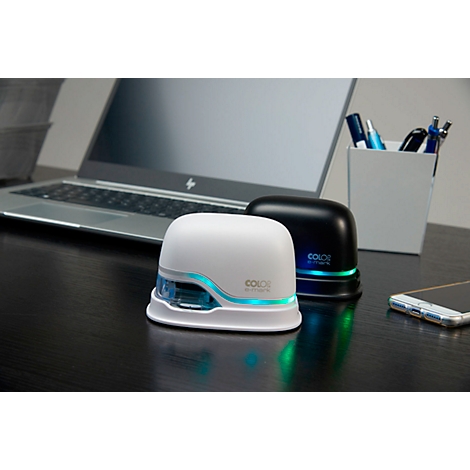 Two stamp devices, white and black, are on a dark desk. Laptop, pens and mobile phone in the background.