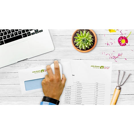 A hand operates the mouse next to a laptop, documents, and a small plant on a white wooden table. A small gardening tool is on the right.
