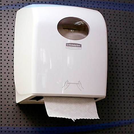 Distributeur d'essuie-mains en papier blanc sur tôle perforée grise. Fenêtre de visualisation ronde en haut, sortie de papier en bas. Inscription : Kimberly Clark Professional.