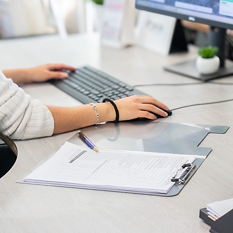 Person arbeitet an einem Schreibtisch mit Tastatur, Maus und einem Klemmbrett.