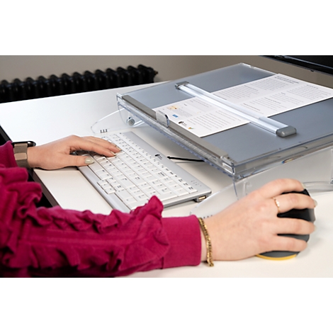Person using keyboard and mouse at a desk. A document lies on a lectern. Arm with a golden bracelet.