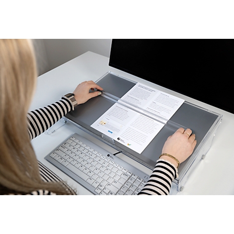 Woman at desk, reading documents placed in a clear holder in front of the monitor.