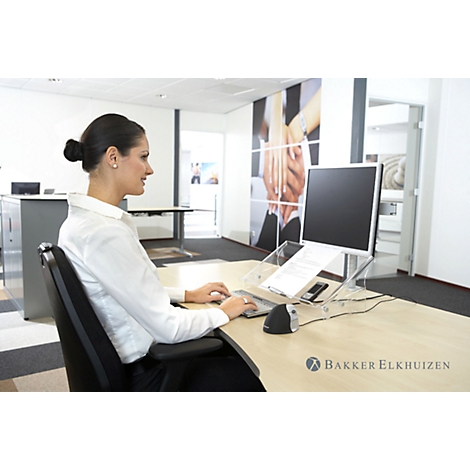 A woman sits at a desk, typing on a keyboard, and looking at a monitor. On the desk is a monitor stand with documents.