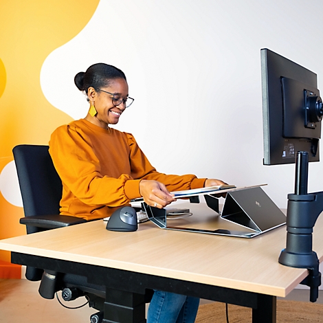A smiling woman in an orange blouse sits at a height-adjustable desk, holding papers and looking at a laptop.