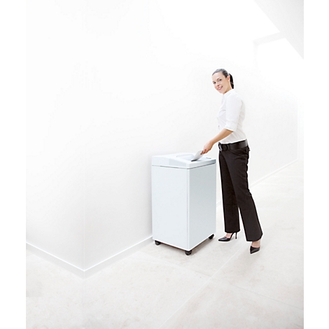 A woman smiles as she puts paper into a white shredder. She wears a white blouse and black pants.