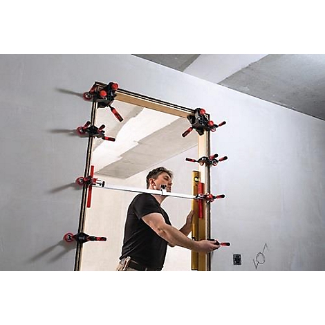 Craftsman installs door frame using clamping device. He uses spirit level and measuring bar. Black T-shirt.