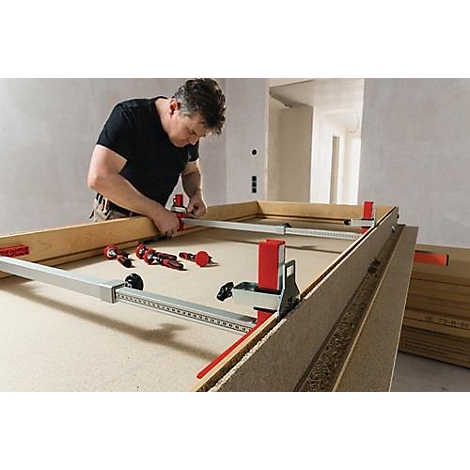 A man builds a wooden frame with tools. Red and silver clamps and tools lie on the surface.