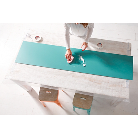 Person wipes on a turquoise surface with a sponge. Underneath a wooden table, next to it two stools.