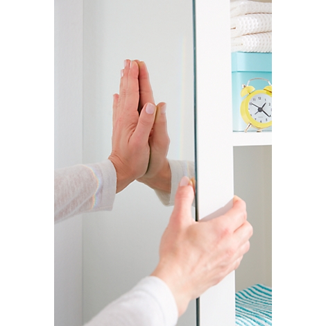 Hands touching a mirrored door located on a white cabinet. In the background there is a shelf with towels, a box and a clock.
