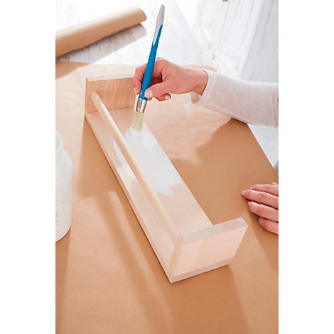 A hand paints a wooden shelf with white paint. The almost finished shelf is lying on brown paper.