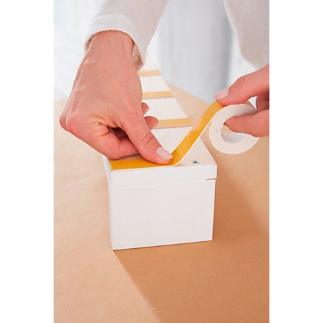 Person is attaching yellow tape to a white box. A roll of tape is ready.