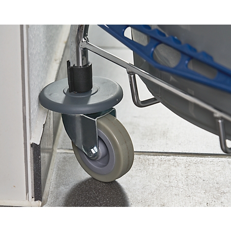 Close-up of a cart wheel standing against a wall. The wheel is grey, the bracket is silver. The surroundings are grey and white.