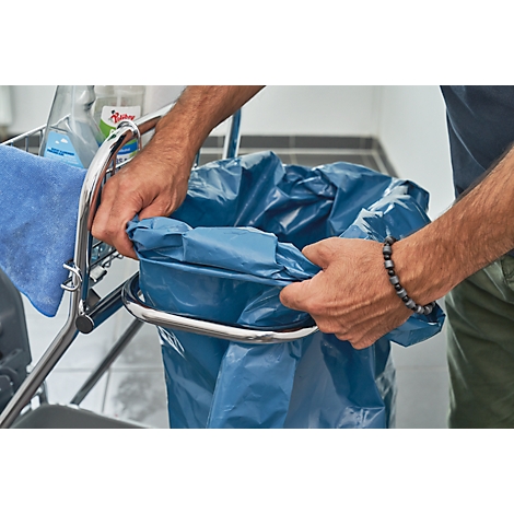 Man fixing a blue trash bag in a trash can. In the background a cart with towel and cleaning products.