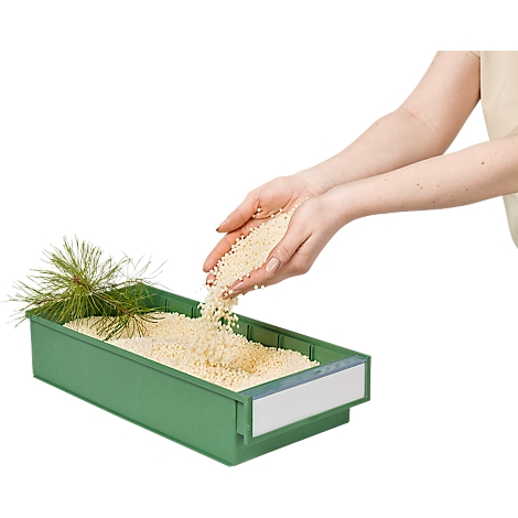 Hands pouring light-colored granules into a green bin. A pine needle lies next to it.