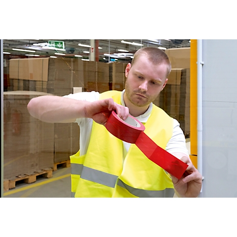 A man in a yellow vest is sticking red tape. In the background pallets and boxes.