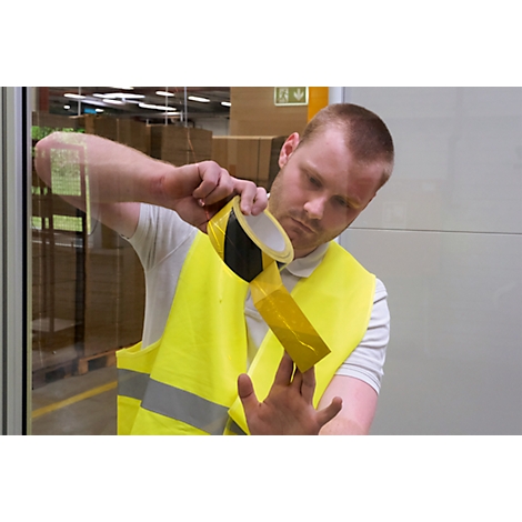 A man is applying yellow warning tape to a yellow safety vest. He is holding the roll and the vest. In the background a warehouse.