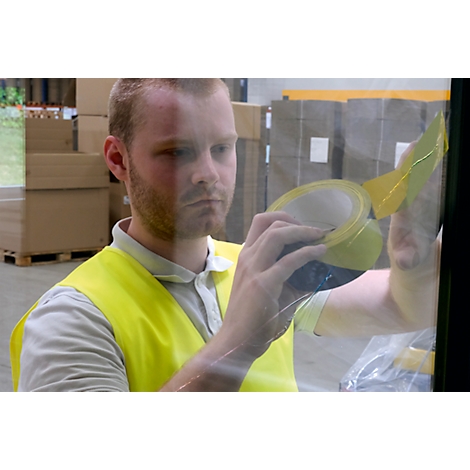 A man in a yellow vest is taping a window with tape. Boxes and pallets can be seen in the background.