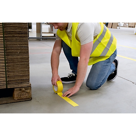 A man kneels and applies yellow tape to the concrete floor. He wears a yellow safety vest.