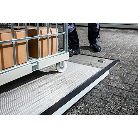 A worker pushes a trolley with boxes over a ramp. Paving stones on the right.