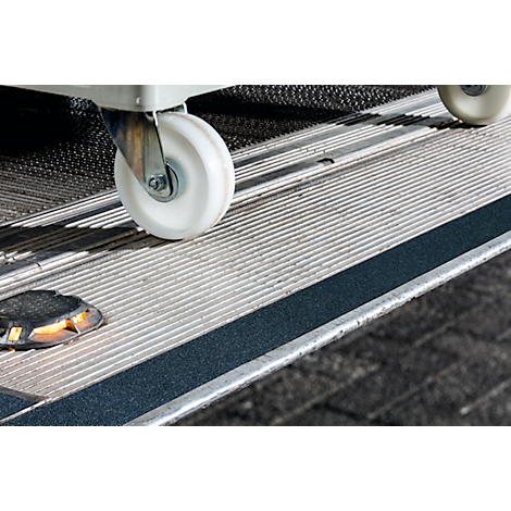 Close-up of a wheelchair ramp featuring a white wheel and a black non-slip strip. The surface is made of corrugated metal.