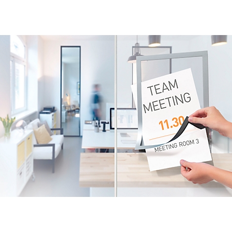 Hands lift a note saying "Team Meeting 11.30 Meeting Room 3" in front of a glass office wall.